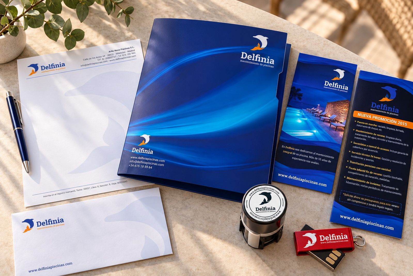 Papelería de Delfinia sobre una mesa de piedra: folio, sobre, carpeta, folleto, sello y memoria USB